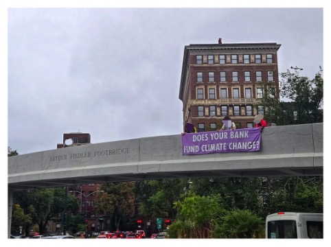 A photograph of a bridge over an urban street. A few people on the bridge hold a purple banner with the words DOES YOUR BANK FUND CLIMATE CHANGE?