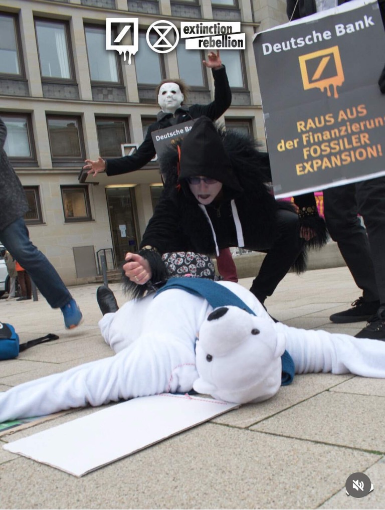 Uma foto de uma cena de teatro de rua num espaço urbano ao arlivre. Alguém vestido de urso polar está deitado no chão, aparentemente aser esfaqueado no peito por uma personagem com pele preta. Outro artistasegura um cartaz que diz "Deutsche Bank raus aus der finanzieruns fossilerexpansion!"