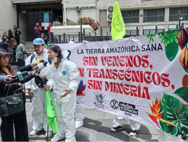 A photo of two people speaking into microphones. They stand beside and in front of a large banner that reads "la tierra Amazonica sana sin venenos sin transgenicos, sin mineria".