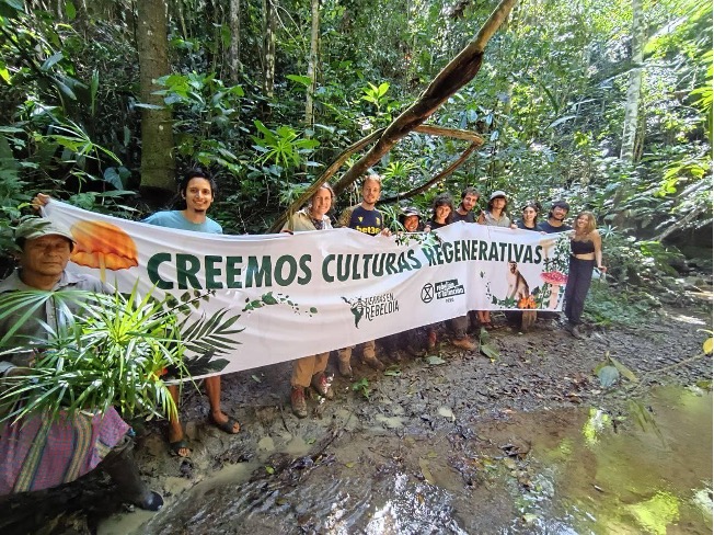 A photo of a group of people holding a long banner in front of a tropical forest edge. The banner reads "creemos culturas regenertivas".