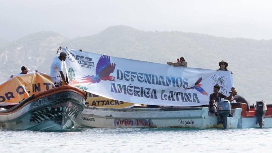 A photograph evidently of the same boats and people as the previous photo, but from a different angle and closer-up. The people are holding up a white banner that reads "Defendamos America Latina".