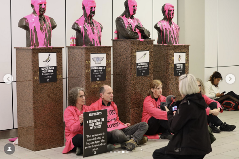 Photo of four bronze busts covered in dripping pink paint. Protesters wearing pink jackets sit on the floor holding signs.